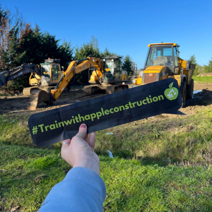 A sign that reads '# train with apple construction' being held up in front of some construction machinery.